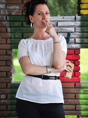 Fully dressed dark brown Mina smokes a cigarette in front of a brick wall